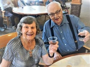 Elderly man in blue shirt and black overalls and an elderly woman in a grey dress holding a glass of red wine in a cheers
