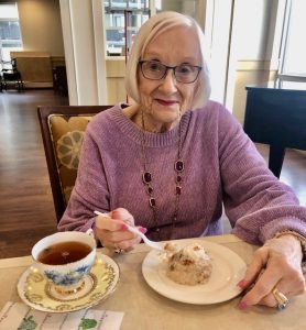 Youthful 90 year old woman with long hair and glasses, pink sweather, having high tea.