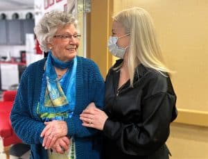 Older lady with short hair and glasses, wearing a blue patterned silk scarf looking at a young woman with long blond hair; elbows linked and both smiling.