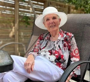 Senior woman in white had and pants, flower patterned shirt, sitting on a patio with legs crossed