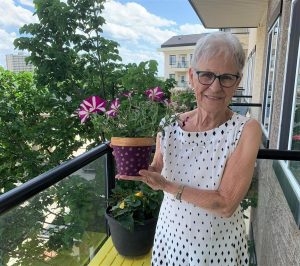 Woman with short grey hair wearing a sundress; holding a planter with flower on her balcony at Sturgeon Creek I Retirement Residence