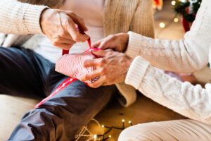 Unrecognizable senior couple in white woolen sweaters sitting on the floor, wrapping Christmas gifts together.