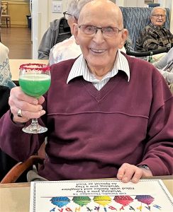 Older senior in burgundy sweater and white collar shirt holding a margarita cocktail, happy birthday placemat in front of him.
