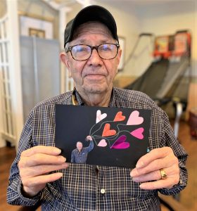 Elderly man in glasses and baseball cap holding a heart craft card