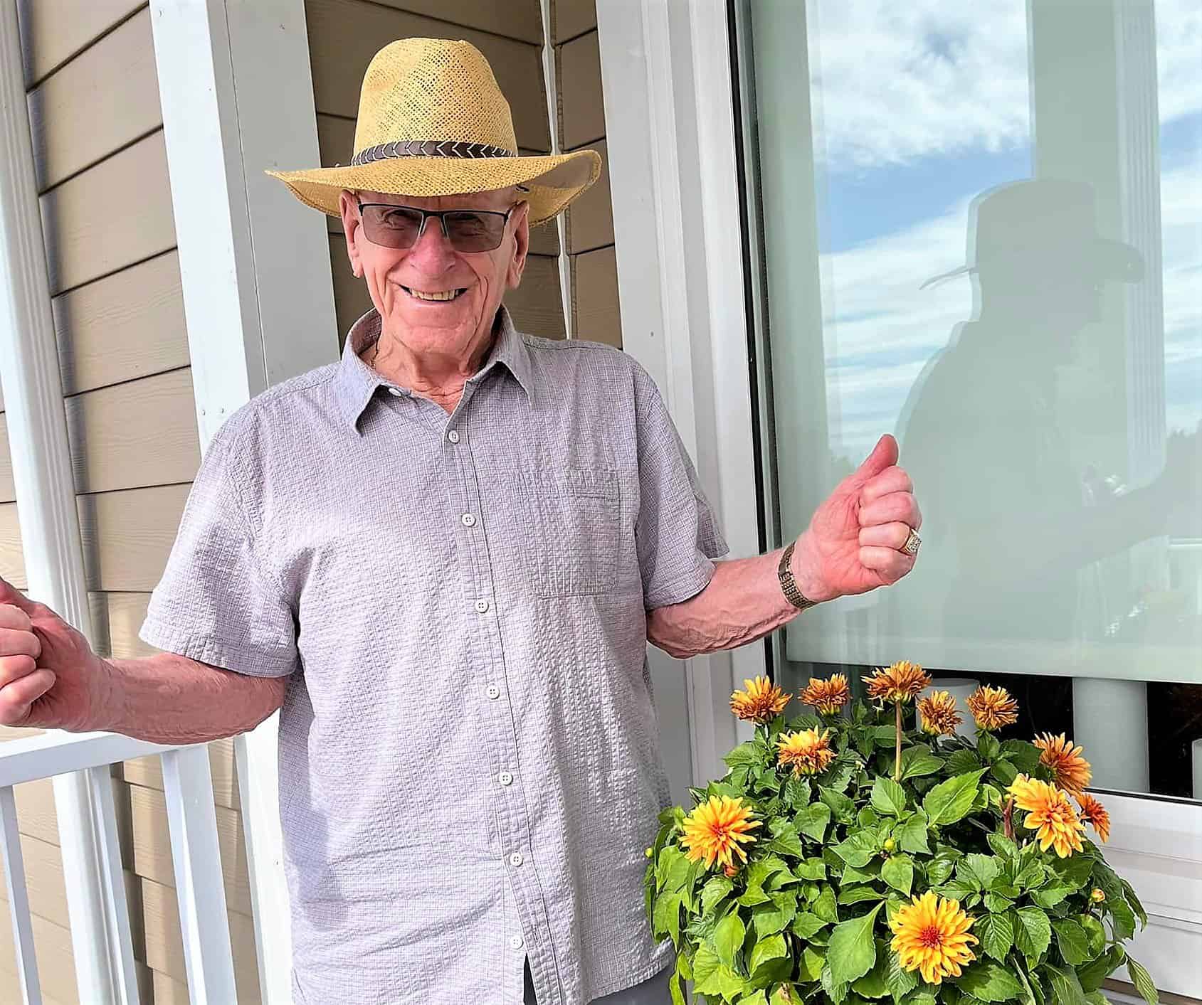 Senior man win straw hat, sunglasses, short sleeve shirt giving the thumbs up; balcony; summer container garden.