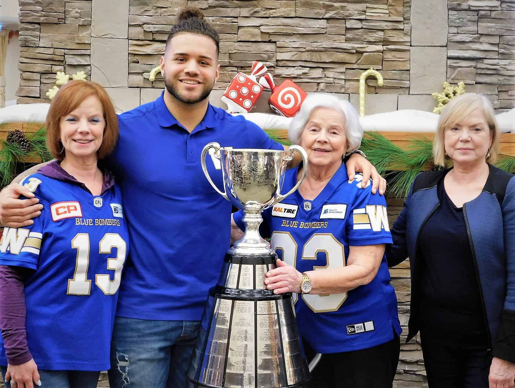 Head-To-Head: Which Retirement Residence Has the Grey Cup Edge? - All Seniors Care NFL player standing with seniors and the Grey Cup in front of a fireplace.