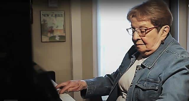 Older woman wearing glasses and blue jacket playing the piano.
