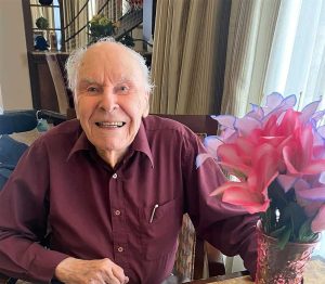 Oral Health Tips for Seniors and Caregivers - All Seniors Care Senior man, smiling, balding, in a burgundy shirt, sitting next to a bouquet of pink flowers.