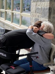 Journée mondiale Alzheimer: Comment soutenir les personnes atteintes de démence - All Seniors Care Caregiver in black bending down to hug older woman with dementia, siting with a walker